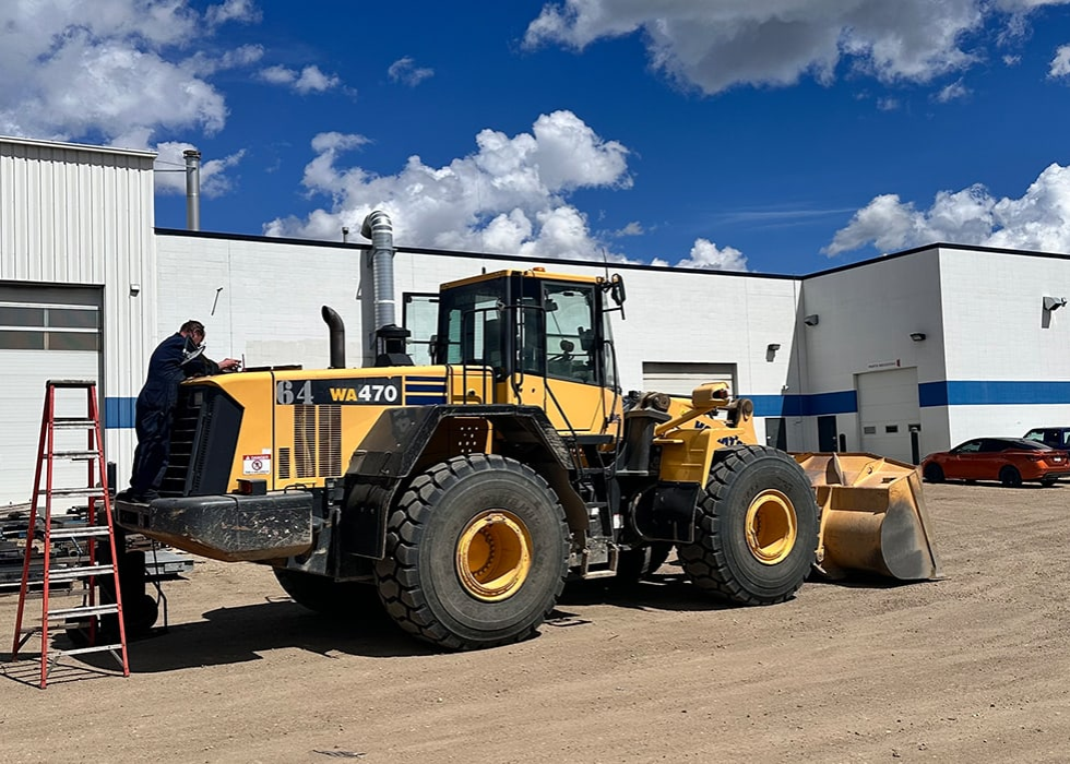 A large yellow vehicle is parked on a sandy surface near a building