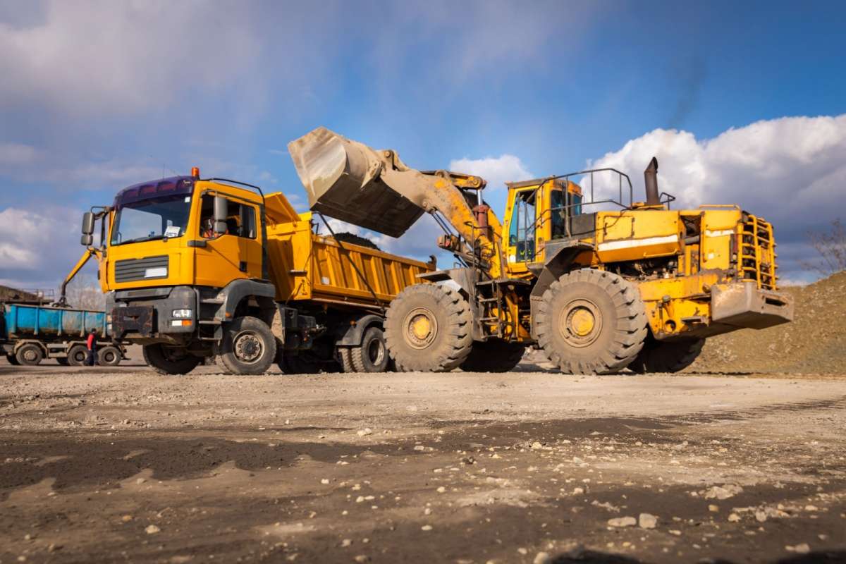 A large wheel loader lifting and dumping soil into a yellow dump truck at an outdoor construction site under a partly cloudy sky.