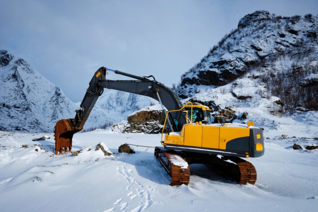 Yellow excavator operating in deep snow with snow-covered mountains in background, demonstrating heavy equipment requiring proper cooling system winterization.