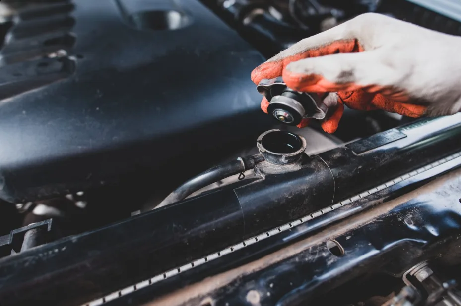 A hand with orange gloves tests the radiator cap on a car engine