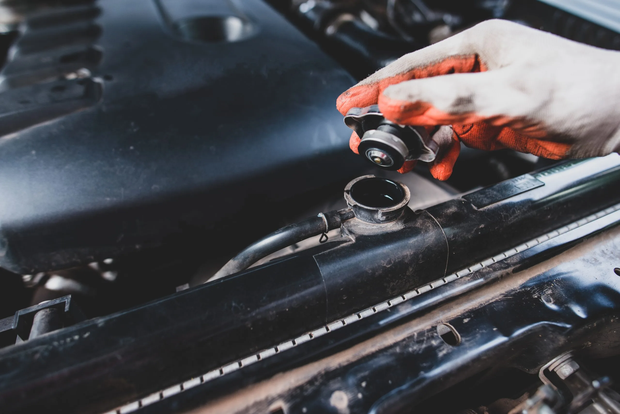 A hand with orange gloves is removing a cap from a car radiator